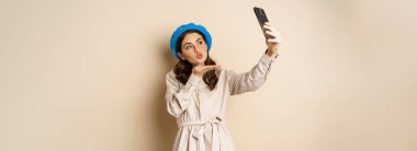 Beautiful modern woman posing for selfie, holding smartphone, taking picture photo on mobile phone, standing happy against beige background.