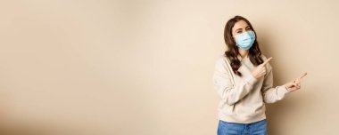 Happy smiling caucasian woman wearing medical face mask from coronavirus pandemic, pointing fingers right, showing advertisement, beige background.