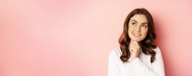 Close up portrait of coquettish smiling woman, glamour girl thinking, looking thoughtful, standing over pink background.