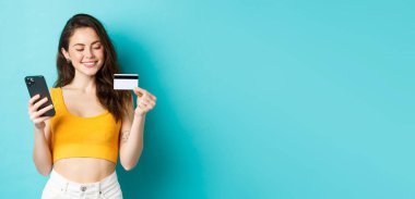 Plastik kredi kartlı şık bir kız, gülümsüyor, online siparişi ödüyor, akıllı telefondan alışveriş yapıyor, mavi arka planda duruyor. Boşluğu kopyala