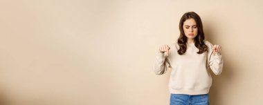 Sad and disappointed girl pointing, looking down with upset grimace moody face, standing over beige background.