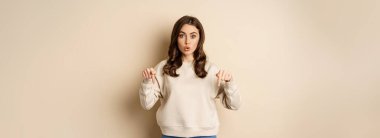 Enthusiastic attractive girl pointing fingers down, showing advertisement, link or promo below, standing over beige background.