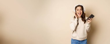 Smiling happy woman listening music in headphones on smartphone, laughing excited, standing over beige background.