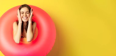Happy beautiful young woman relaxing on summer vacation, wearing pink swim ring for beach holiday, standing against yellow background.