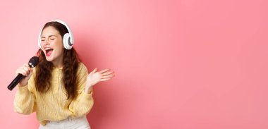 Karaoke yapan genç bir kadının portresi, kulaklık takan, şarkı söyleyen, mikrofonu tutan, pembe arka planda duran.