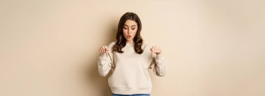 Surprised girl pointing and looking down at smth curious below, beige background.