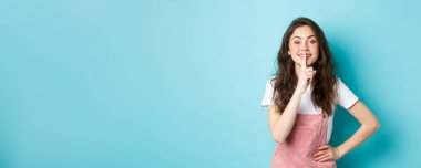 Sessiz ol. Şirin esmer kız susuyor ve gülümsüyor, bir sırrı var, parmaklarını ağzında tutuyor ve kameraya nazik bakıyor, mavi arka planda duruyor..