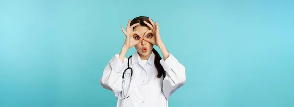 面白いセラピスト 女の子の医者は大丈夫 双眼鏡ゼロジェスチャー目と笑顔 周りをだますと楽しい 青い背景の上に立って — ストック写真
