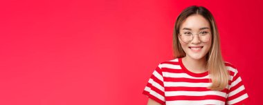 Yakın plan, neşeli Asyalı sarışın kız, gözlük takıyor, gülümsüyor, hoş gülümsüyor, dostça gülümsüyor, arkadaşça sırıtıyor, arkadaşça konuşuyor, mutlu bir şekilde duruyor, olumlu duygular sergiliyor, kırmızı arka planda duruyor..