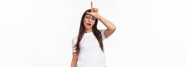 Portrait of arrogant and sassy, confident young stylish woman in glasses, mocking person showing rude loser sign on forehead, laughing at person who lost competition, stand white background.