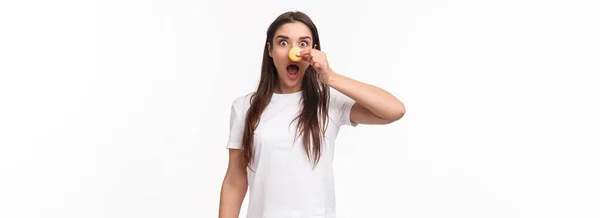面白い愛らしい若いブルネットの女性の肖像Tシャツ 口を開けて興奮し 熱狂的に見える マカロンから鼻を作る冗談とカメラを見つめ おいしいデザートを食べる 白い背景 — ストック写真
