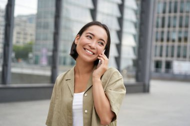 Gülümseyen Koreli genç kız cep telefonuyla konuşuyor ve şehirde yürüyor. Akıllı telefonuyla sokakta poz veren mutlu kadın..