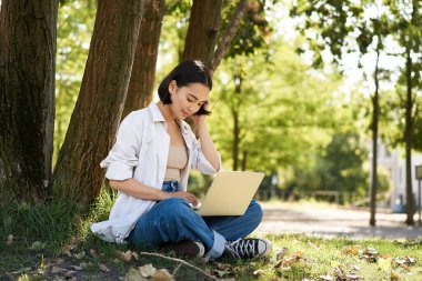 Uzaktan kumanda. Gülümseyen Asyalı kız, uzaktan ödev yapan öğrenci, dizüstü bilgisayarı ağacın yanında oturuyor ve daktilo yazıyor..