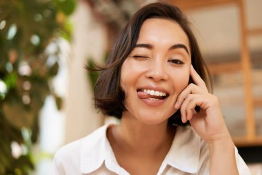 Kafede oturmuş, neşeli bir şekilde sohbet eden, kameraya gülümseyen komik Asyalı genç kadının yakın portresi..