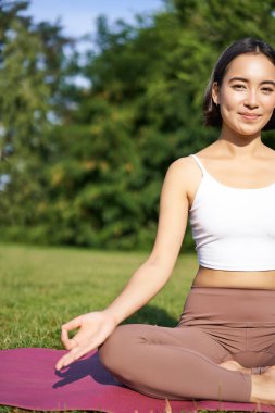 Genç Asyalı kadının spor paspasına oturup meditasyon yaparken, parkta sakinleştirici ve rahatlatıcı egzersizler yaparken, zen işareti gösterirken dikey çekimi..