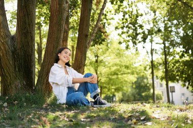 Güzel gülümseyen kız parkta bir ağacın yanında oturur, dışarıda doğanın tadını çıkarır, dinlenir ve temiz havada dinlenir. Boşluğu kopyala