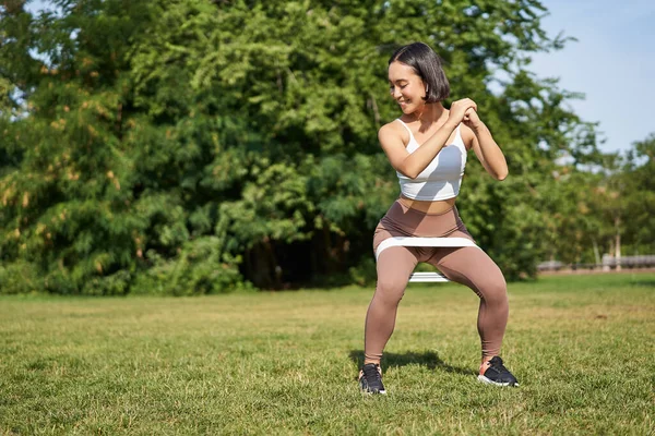 Woman using stretching band, resistance fitness rope for workout in park, doing squats, training on fresh air.
