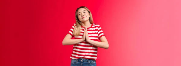 車を貸してください 可愛いですクリニー愚かなアジアのブロンドの女の子は手を祈る保持します 頭の笑顔柔らかいカメラを傾けます 好意を求めます 天使のような赤い背景行為に立つ友人のヘルプ — ストック写真