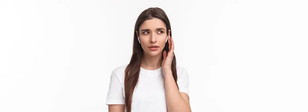 Tシャツを着た若い白人女性のクローズアップ肖像 ワイヤレスヘッドフォンを身に着けて 密接に耳を傾け ボリュームを固定し 電話に答えるためにイヤホンに触れ 音楽を楽しんで白い背景に立って — ストック写真