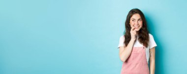 Şirin kıvırcık saçlı düşünceli bir kız, parmağını ısırıyor ve reklam logosuna bakıyor, gülümsüyor bir şeyler denemeyi düşünüyor, mavi arka planda duruyor..