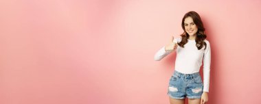 Attractive young woman showing thumbs up in approval, like and agree, satisfied with quality, recommending store or beauty salon, pink spring background.