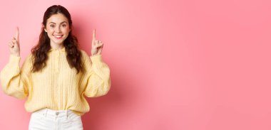 Çekici beyaz bir kadının, reklamını gösterip, kameraya gülümsediği, pembe arka planda durduğu bir görüntü..