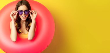 Close up portrait of stylish young woman, girl with swimming ring and summer sunglasses, ready for beach holiday, vacation, standing over yellow background.