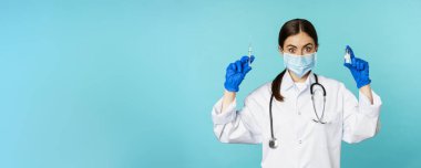 Gülümseyen sağlık personeli, maske takan doktor ve lastik eldiven, mavi arka planda duran covid-19 salgınından şırınga ve aşı gösteriyor..