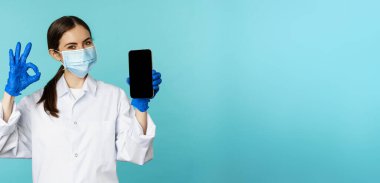 Portrait of doctor in medical face mask and gloves, showing mobile phone app, smartphone screen and okay sign, recommending online checkup website, standing over torquoise background.