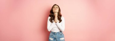 Please. Young stylish girl begging, holding hands in pray, need help, pleading, supplicating, standing against pink background.