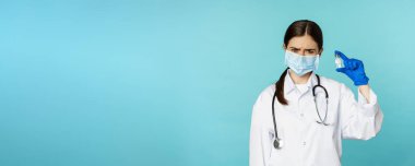 Üzgün ve hayal kırıklığına uğramış maskeli kadın doktor, aşıyı üzgün bir suratla gösteriyor, mavi arka planda üniformalı..