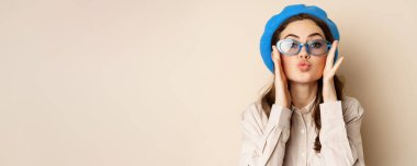 Beautiful stylish woman in sunglasses, blowing air kiss on hands near lips, gazing softly and flirty at camera, posing against beige background.