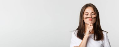 Komik, tatlı esmer kızın yakın çekimi. Surat asıyor, saçlarını şakacı bir şekilde dudağının üzerinde tutuyor..