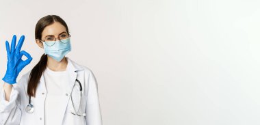 Close up portrait of woman doctor, physician in face mask from coronavirus, showing okay sign in approval, recommending, praise and like smth good, standing over white background.