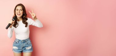 Party girl. Happy young woman singing in microphone, performing song, having fun at event, standing over pink background.