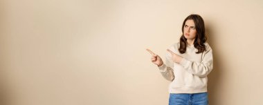 Woman pointing left and staring confused at banner copy space, strange logo, standing over beige background.