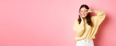 Güzel esmer kadın modelin görüntüsü, gülümseyen ve yüzüne dokunan, göze yakın V işaretini gösteren, mutlu bir şekilde gülen, pembe arka planda duran.