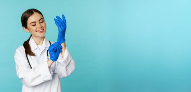 Smiling young woman, medical worker wearing gloves for patient checkup, standing in hospital clinic uniform over blue background.