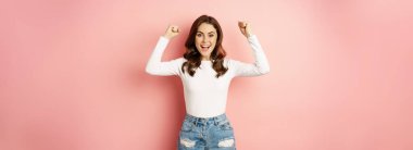 She is a winner. Enthusiastic brunette girl celebrating, jumping from happiness and shouting joyful, achieve goal success, standing over pink background. Victory concept