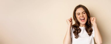 Close up portrait of enthusiastic young woman rejoicing, shouting with joy and satisfaction, celebrating victory, winning and triumphing, standing over beige background.