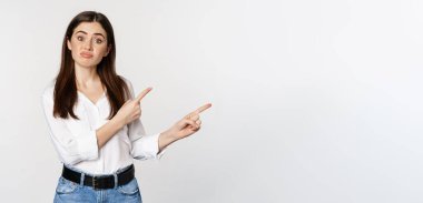 Sad and disappointed cute woman, gloomy girl pointing fingers at logo, complaining, sulking upset, standing in blouse over white background.