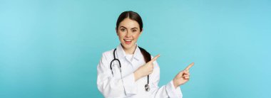 Portrait of smiling medical worker, girl doctor in white coat with stethoscope, pointing fingers left, showing medical clinic advertisement, torquoise background.