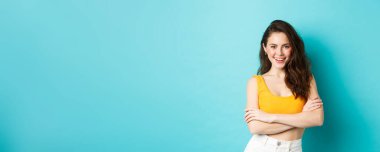 Yaz ve yaşam tarzı konsepti. Kendine güvenen ve küstah çekici bir kadın. Üstü açık giyinmiş kameraya bakıyor. Kollarını göğsüne dayayıp gülümsüyor ve mavi arka planda duruyor..