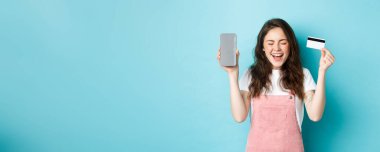 Heyecanlı çekici kadın sevinç çığlığı, boş akıllı telefon ekranı ve plastik kredi kartı gösteriyor, elini kaldırıyor ve gülümsüyor, mavi arka planda duruyor..
