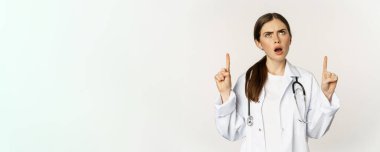 Sad and disappointed female doctor, frowning frustrated, pointing fingers and complaining, standing in white coat over white background.