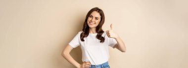 Portrait of modern young woman showing thumbs up, like and approve, smiling pleased, recommending company or website, standing over beige background.