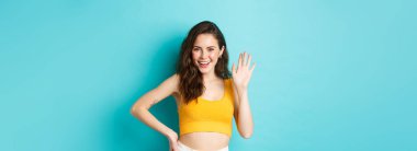 Yaz tatili ve duygu konsepti. Mükemmel vücutlu neşeli arkadaş canlısı kadın, el sallıyor ve merhaba diyor, selam veriyor, selam veriyor, seni selamlıyor, mavi arka planda duruyor..