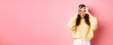 Komik ve tatlı genç bir kadın dudaklarını büzüyor, komik suratlar yapıyor, el dürbünü ve şaşı gözlerle bakıyor, pembe arka planda duruyor..