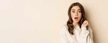 Close up shot of excited woman looking amazed with opened mouth, standing over beige background. Copy space