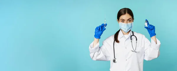 Smiling medical staff, doctor in face mask and rubber gloves, showing syringe and vaccine from covid-19 pandemic, standing over blue background.